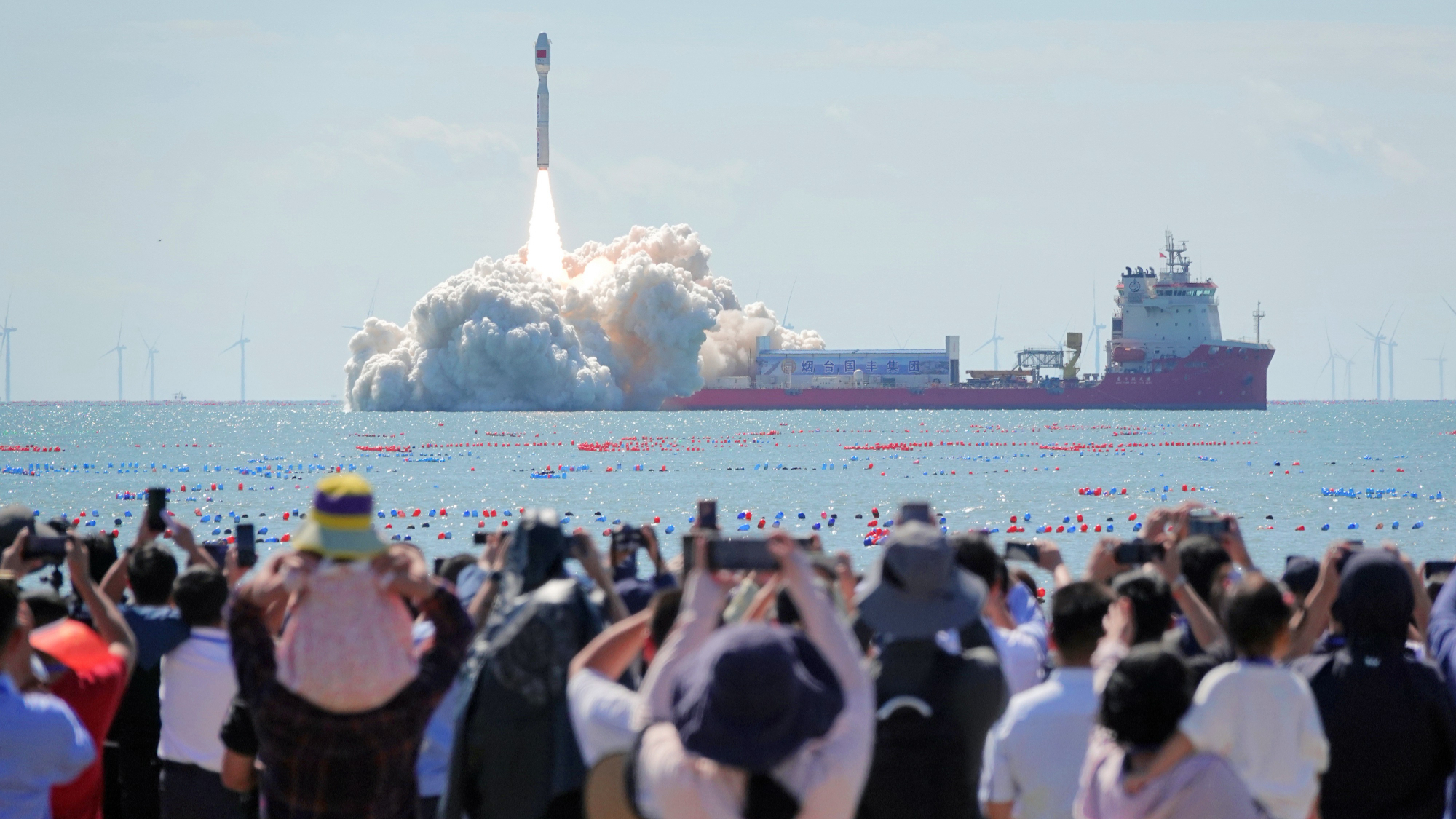 spectators watch a white rocket launch into a blue sky from a ship anchored at sea, not far offshore