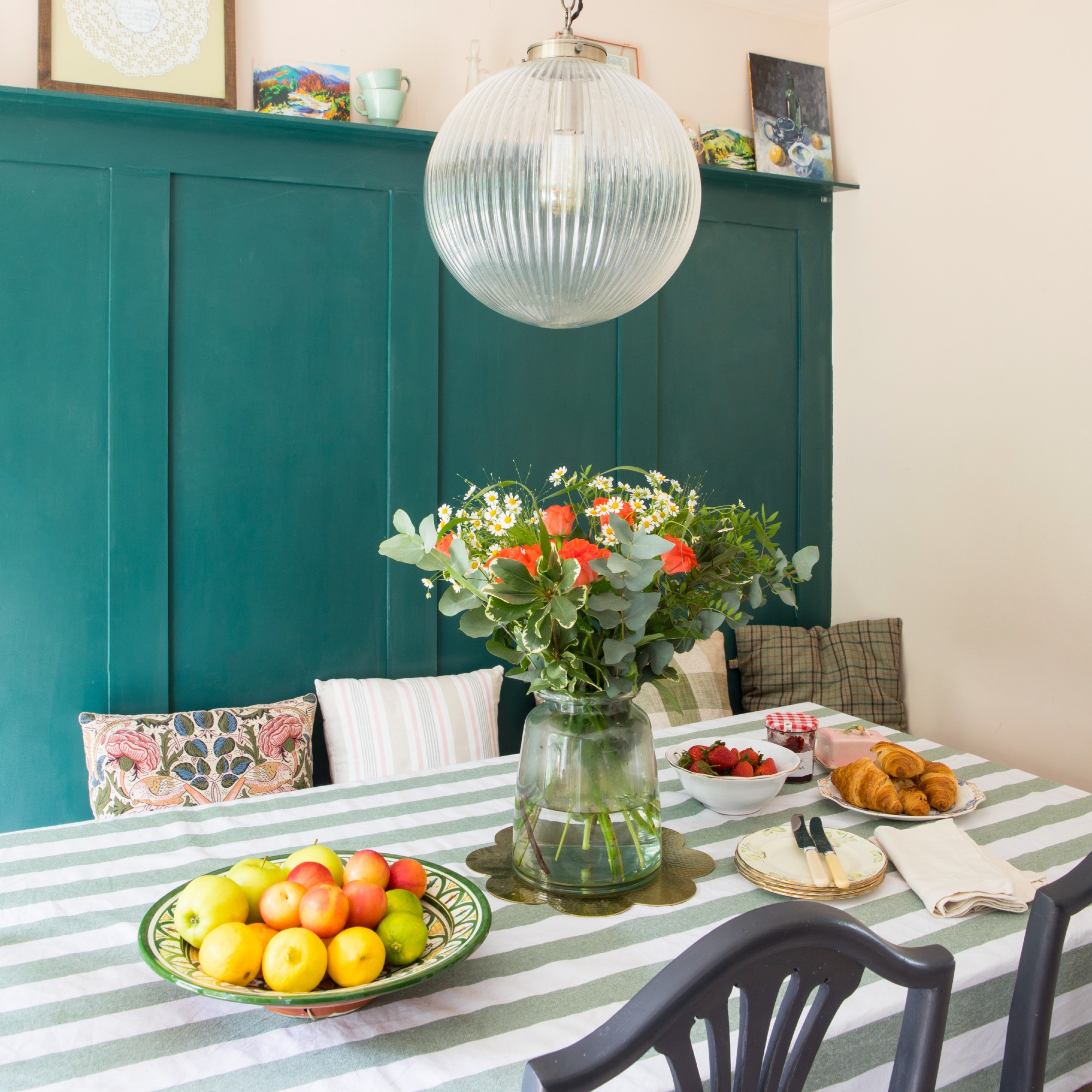A dining table covered with a striped green tablecloth, with a vintage bowl of fruit and a glass vase of flowers on top