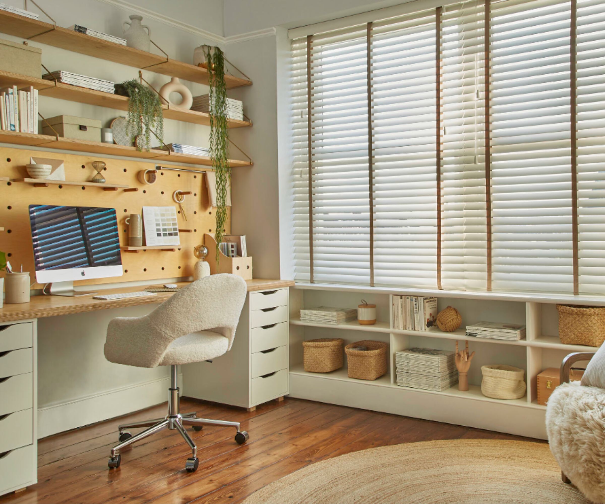 Warm and inviting home office with a wooden pegboard desk, floating shelves, soft swivel chair, and low built-in storage beneath wide windows with blinds.