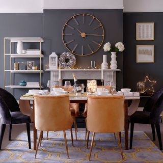 A grey dining room with orange and black velvet chairs, a large wall clock, marble fireplace and patterned rug