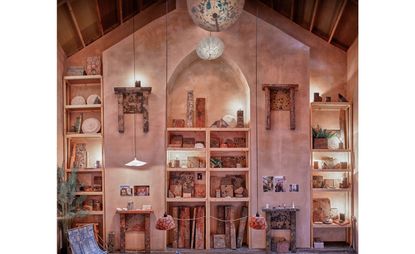 Granby Workshop - arch shelving units on a neutral stone wall, lighting, colourful ornaments, potted plant, small tables with ornaments, wooden slat ceiling with hanging ceiling lights 