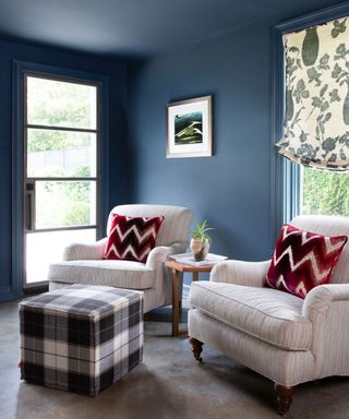 sitting area with dark blue walls and ceiling, beige set of armchairs, plaid footstool and red patterned cushions on chairs