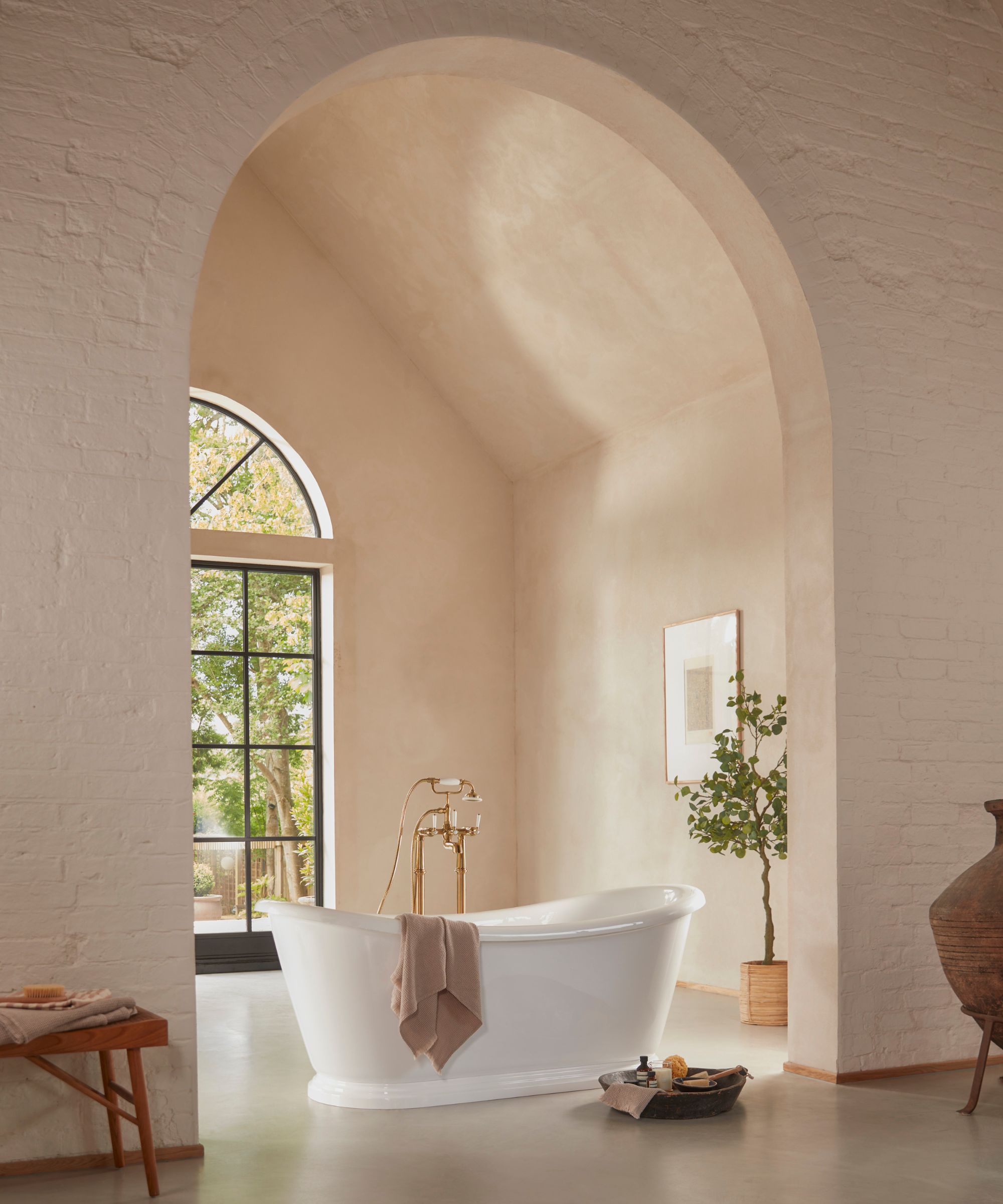 A serene bathroom with a freestanding white bath set beneath a soft arched ceiling, warm plaster walls, brass taps and natural light from a tall window.