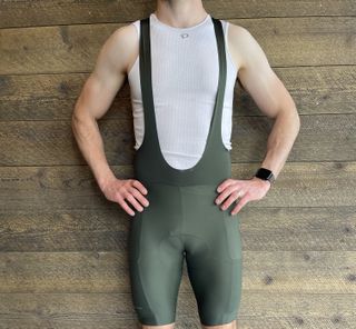 male cyclist standing infront of a wood clad wall modeling the pearl izumi expedition bib short with 2 side pockets