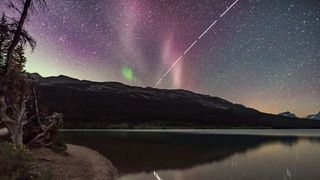 a white streak across the night sky is the ISS in this long exposure photo. A body of water below reflects the cosmic scene and purple and green aurora shine in the sky above.
