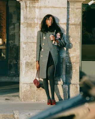 Woman standing in front of Paris wall, wearing gray military coat, tights, and red shoes.