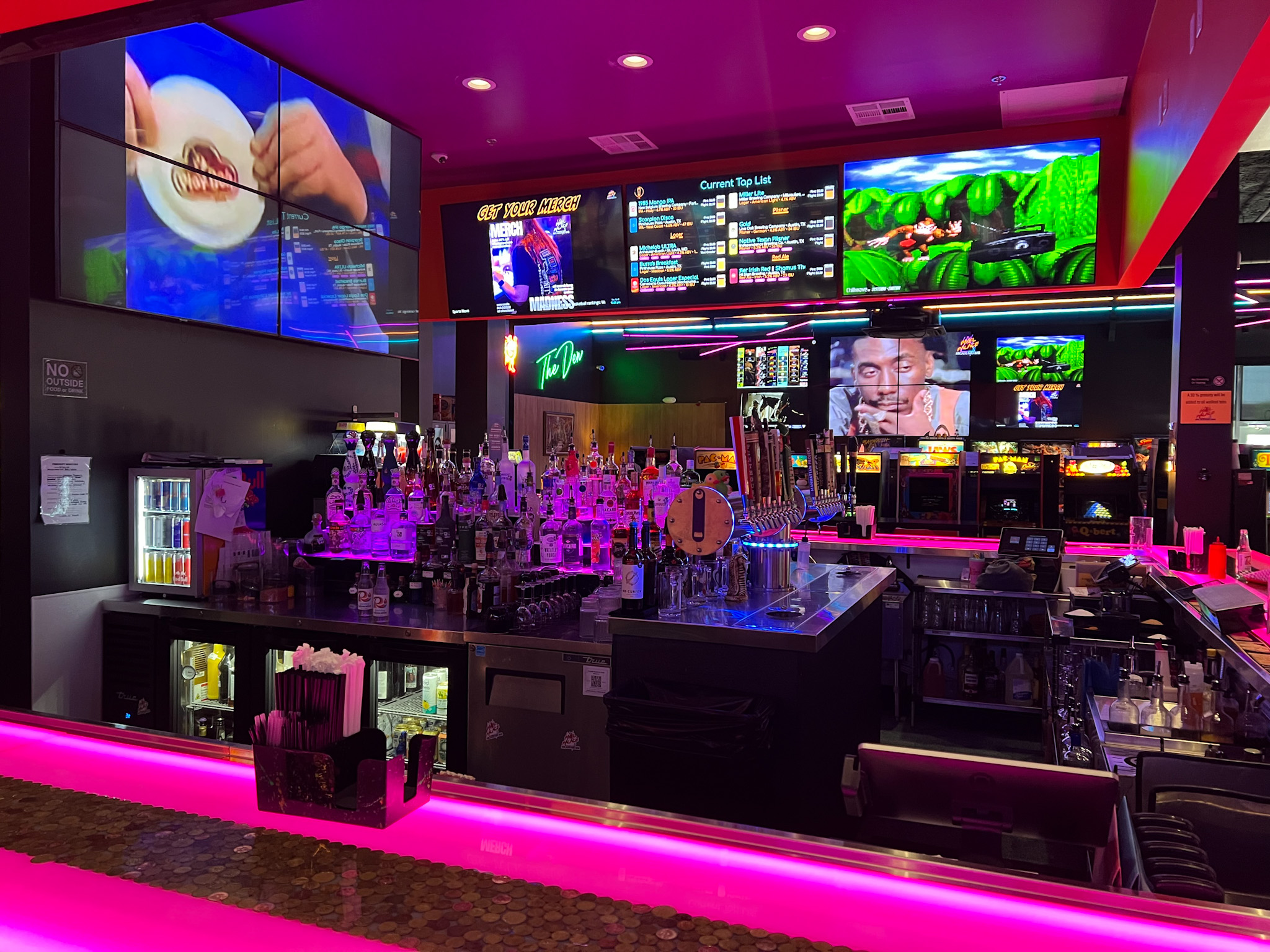 The interior of Pixel Palace arcade and bar showing video games and a bar under bright neon pink lighting.