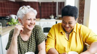 Two female neighbours drinking coffee together in kitchen having fun sharing gossip