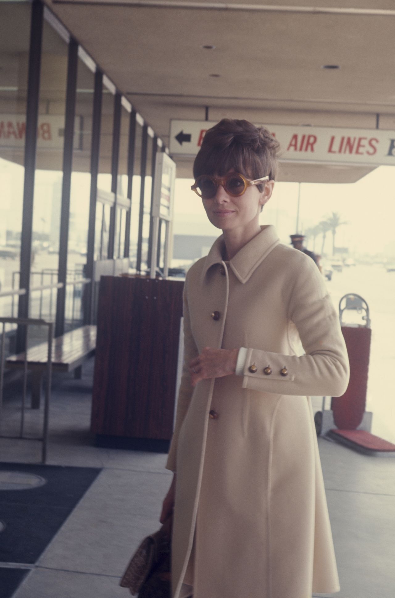 Audrey Hepburn entering an airport dressed in a beige coat with large buttons