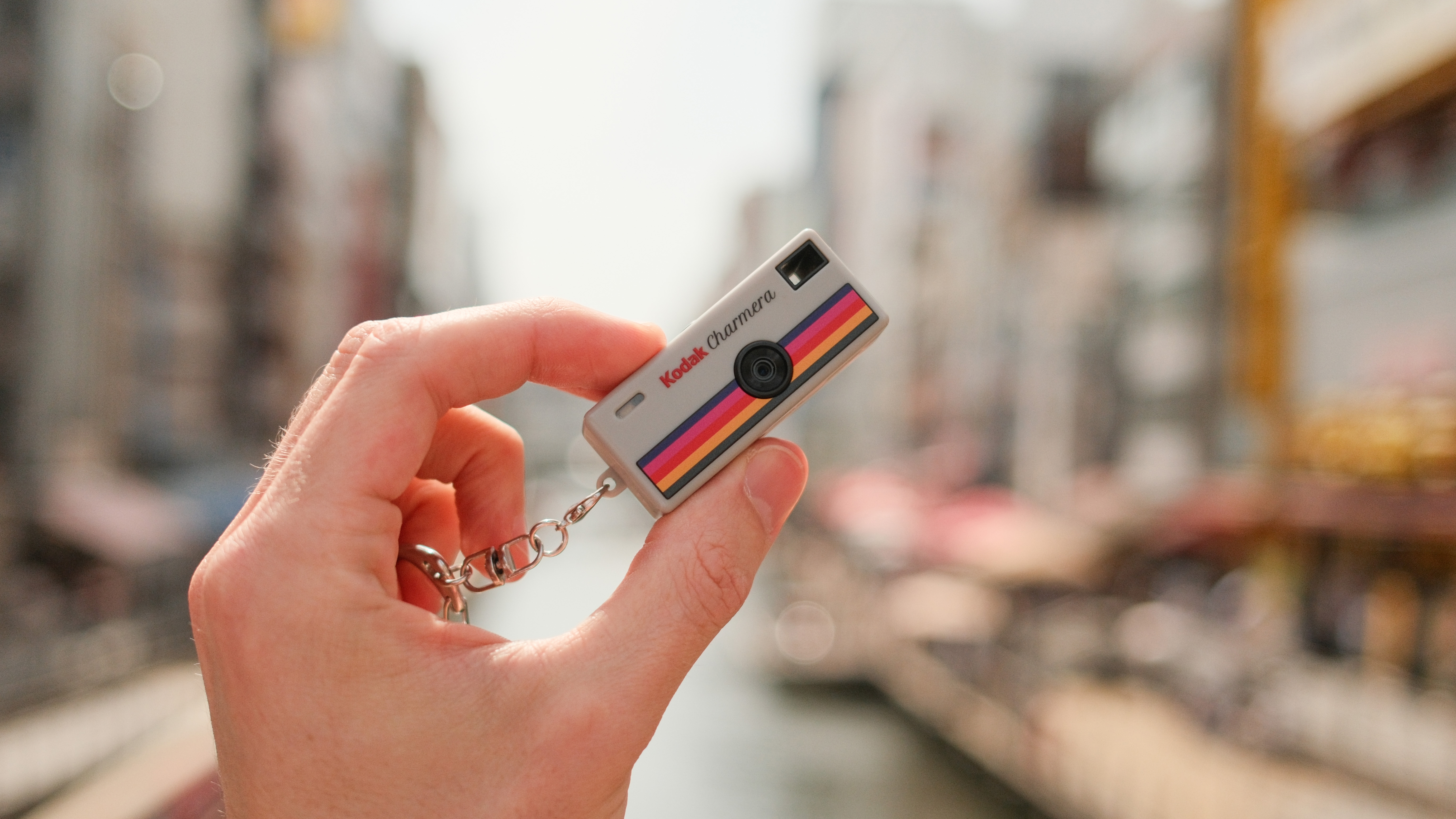 Hand holding a tiny Kodak Charmera keychain camera against a blurred canal background.