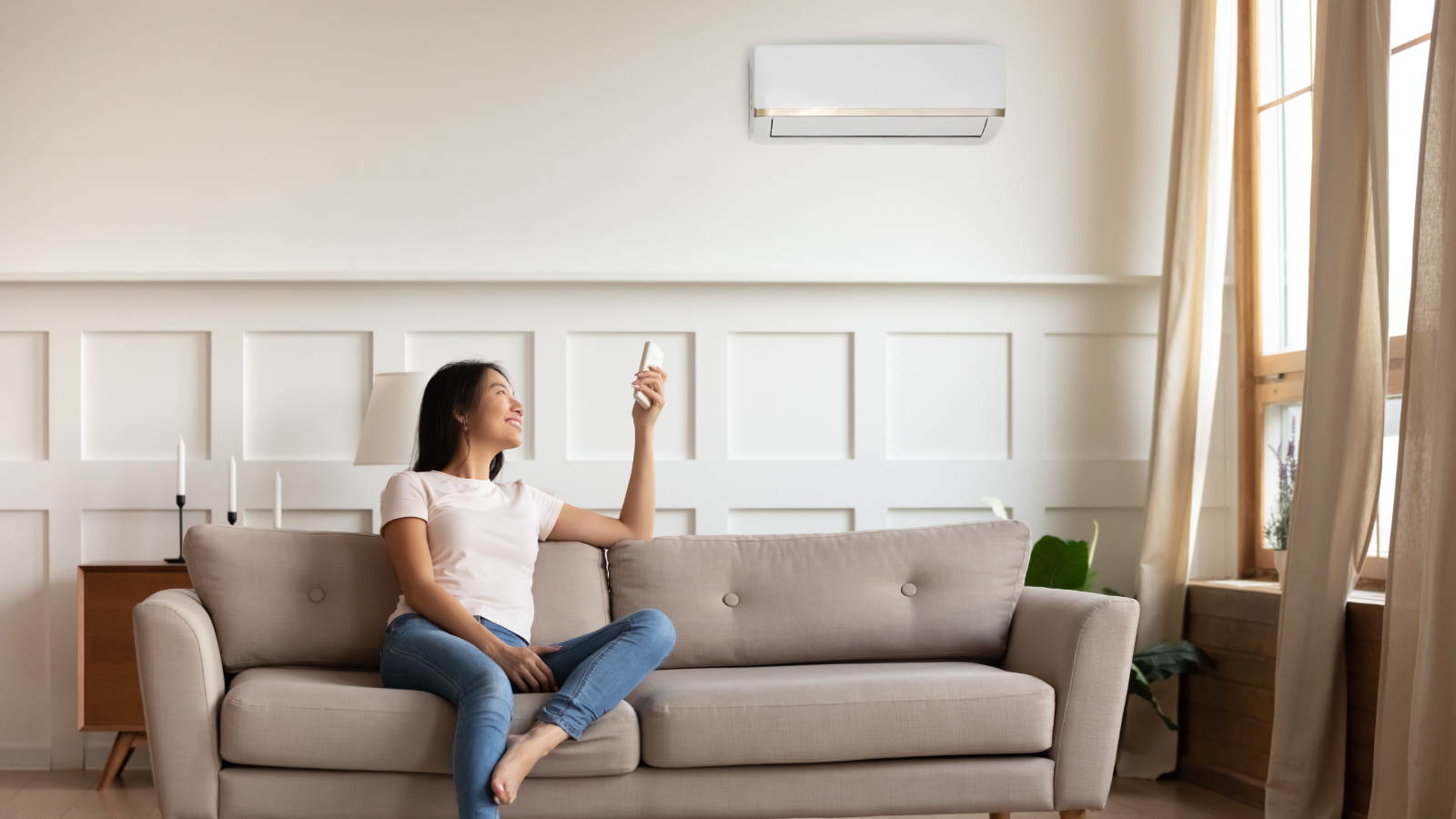 asian woman sitting on sofa in living room pointing remote at air con unit on wall