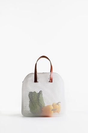 White net bag with brown handles containing green vegetable and yellow peppers. Photographed against a white background