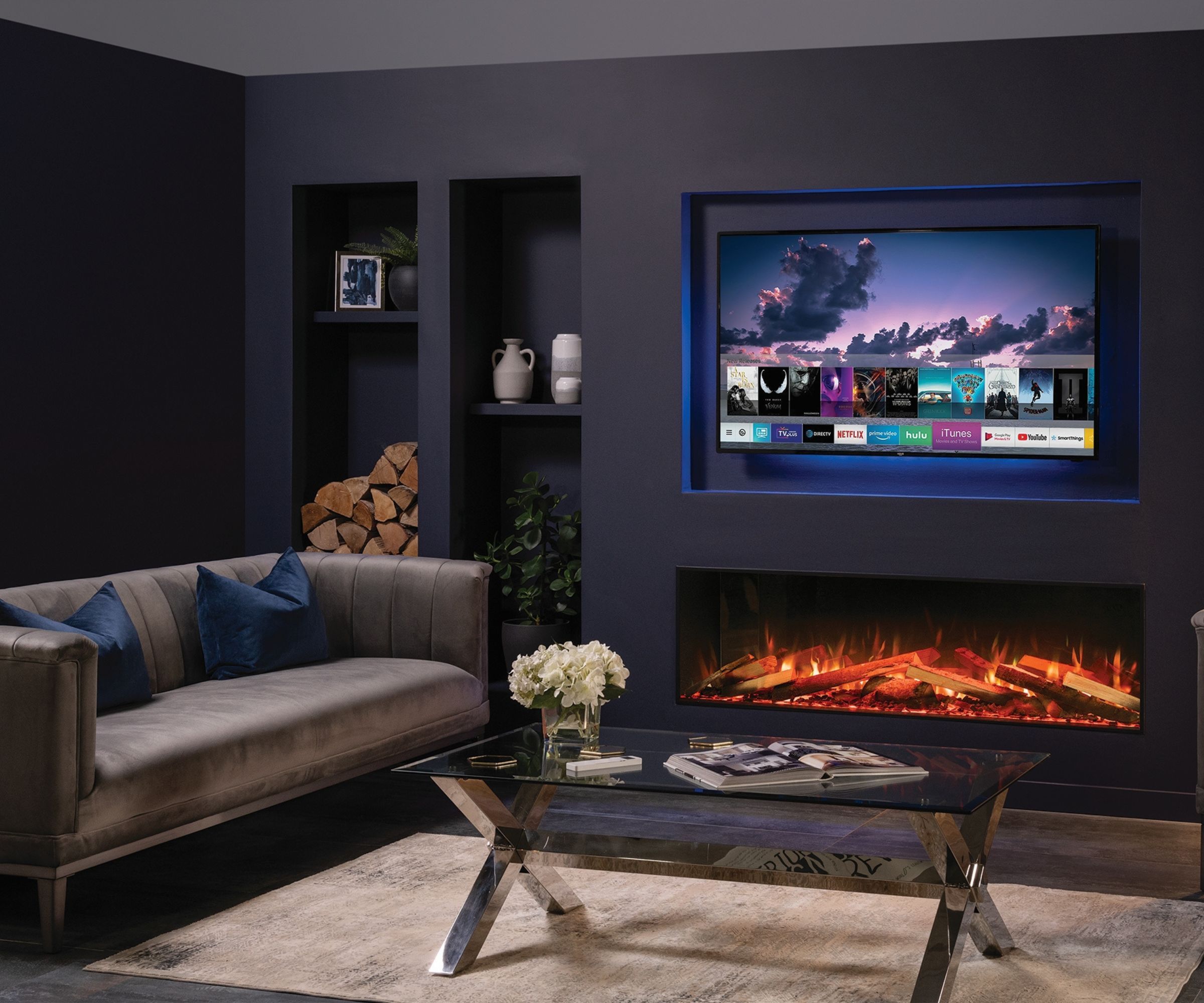 A stylish dark-toned living room with a recessed electric fireplace and wall-mounted TV, navy feature wall with built-in shelving, a grey velvet sofa with blue cushions, and a glass coffee table.