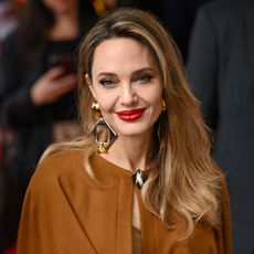 Angelina Jolie smiles wearing large gold earrings and a brown cape