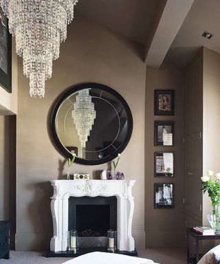 An example of bedroom chandelier ideas showing a bedroom with a large tiered chandelier, circular mirror and white fireplace