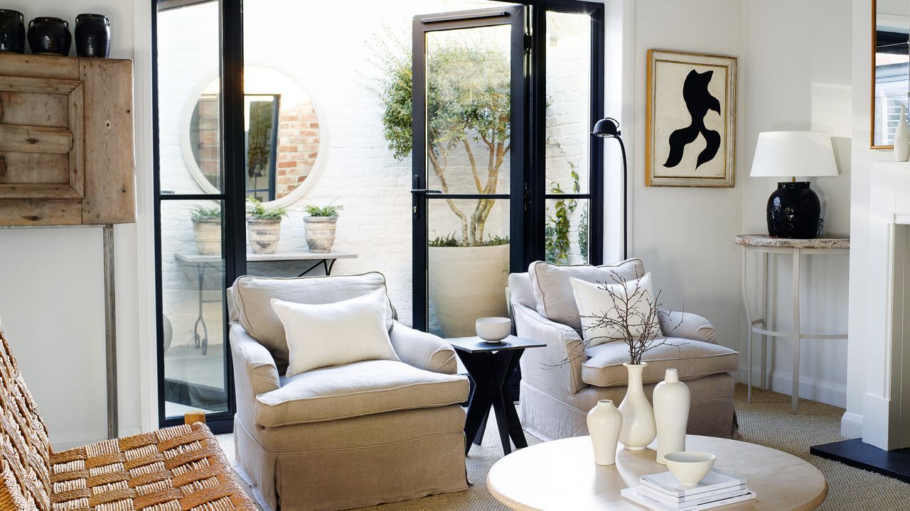 monochrome living room with two linen armchairs, 1970s wooden coffee table, rattan chair and black metal framed doors