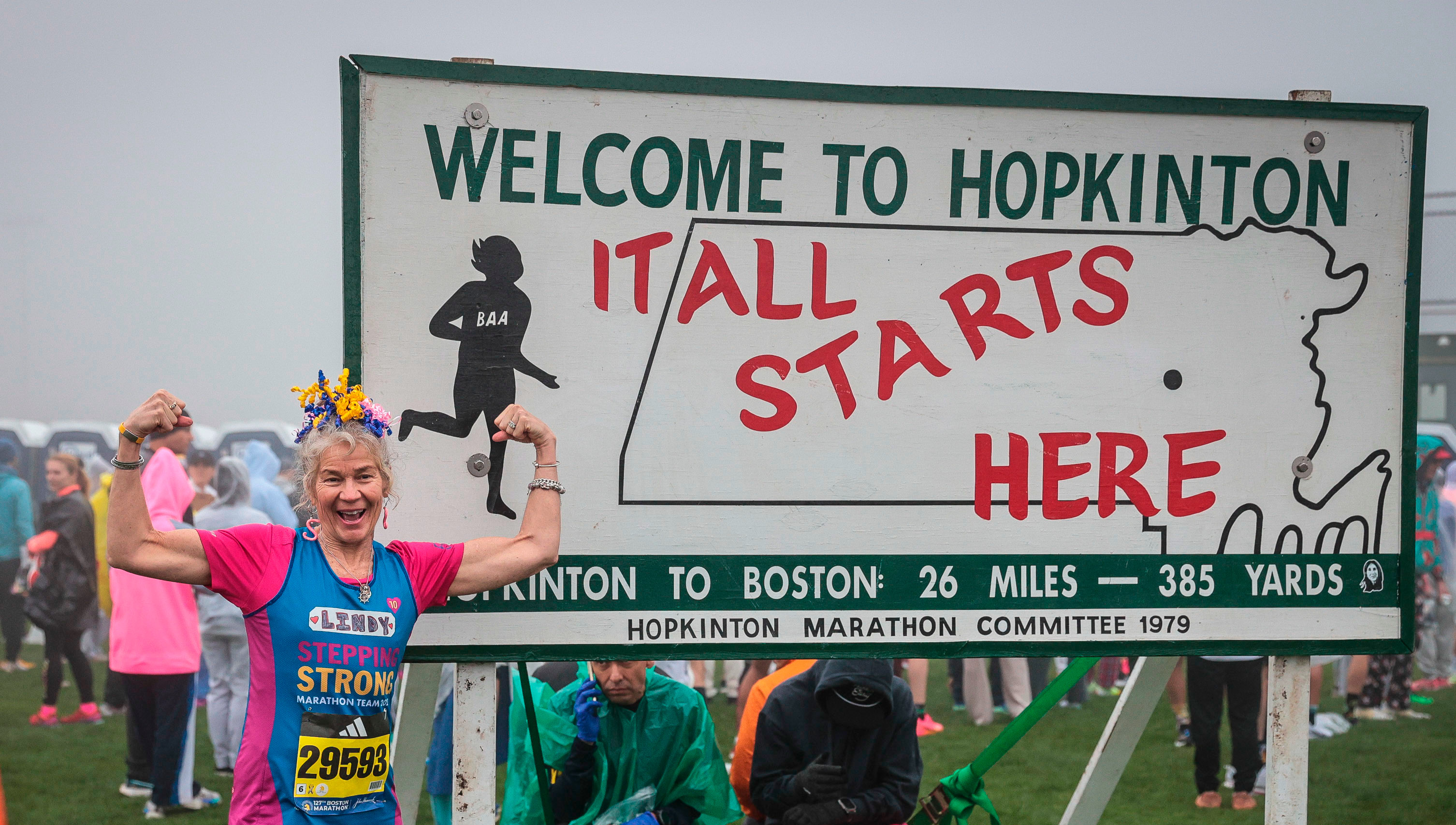 A female runner poses for a picture in front of a sign which reads: Welcome To Hopkinton It All Starts Here Hopkinton To Boston 26 Miles - 385 Yards Hopkinton Marathon Committee 1979