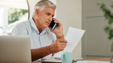 An older man looks at paperwork while talking on the phone.