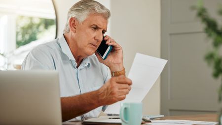 An older man looks at paperwork while talking on the phone.
