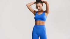 Woman standing with hands behind head in blue workout gear against grey background