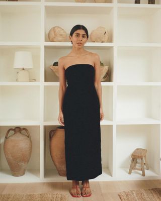 British fashion influencer Monikh Dale poses in her London home in front of white shelves with pottery wearing a black strapless dress and strappy red sandals