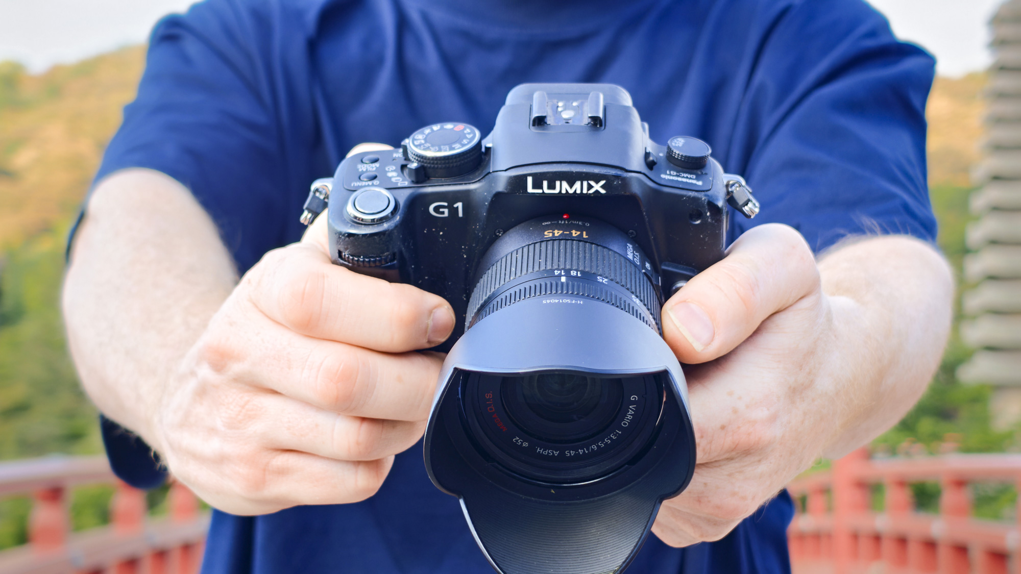 Man holding the Panasonic Lumix G1 mirrorless camera in a rural Japan setting, stood by a red bridge