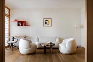 Interior view of the Take suite at the Shinmonzen hotel featuring white walls, wood flooring, wall art, a floor lamp, a red wall-mounted shelving unit, a wooden coffee table with mugs and a teapot on top and a desk. There is also an ‘Akron’ desk chair and an ‘Orbit’ sofa and armchairs, all by Toan Nguyen