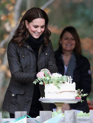 Kate Middleton wearing a brown coat and looking at a white cake trimmed with flowers