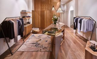 A clothing store with clothing displayed on rails and shelves along the walls, wooden floors, wooden cupboards and a glass topped angled console in the middle of the room.