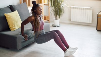 Young black athletic woman doing triceps dips while leaning on the sofa at home