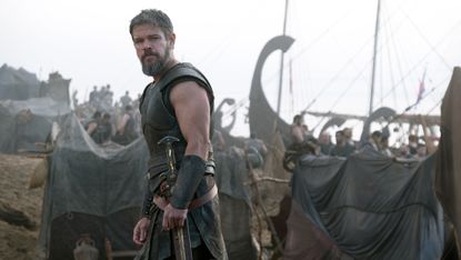 Matt Damon's Odysseus looking over his left shoulder with some ships in the background in The Odyssey