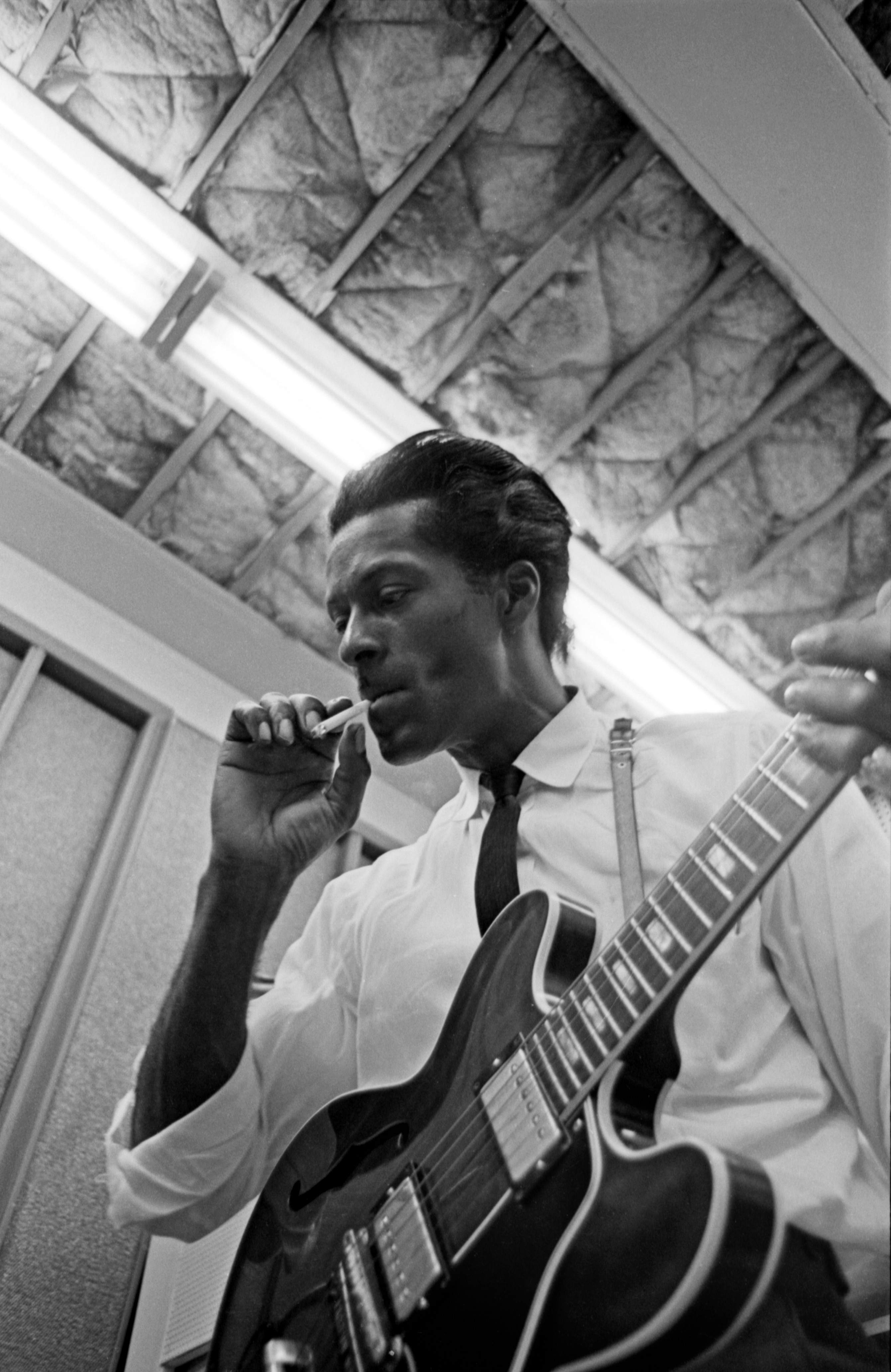 A black and white, low-angle shot of Chuck Berry playing an electric guitar while holding a cigarette in his mouth.