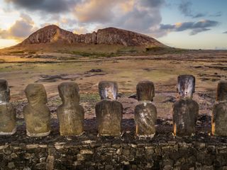 a row of moai statues from behind gaze at the large volcanic structure in the distance.