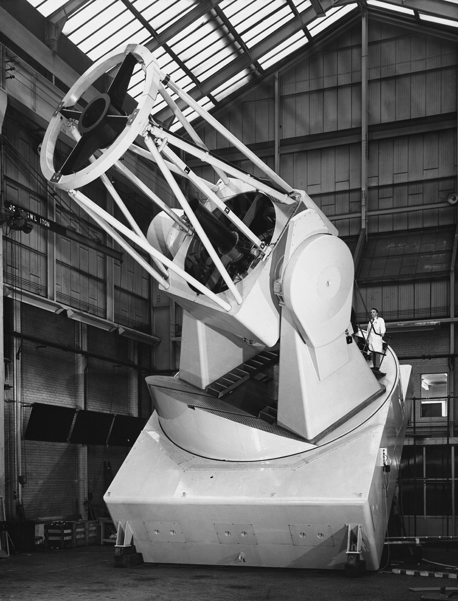 a scientist in a lab coat stands atop a massive telescope towering over him inside a hangar