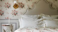 A neutral floral bedroom with statement headboard, white pillowcases, floral bedding, grey wall panelling, white and pink floral wallpaper, white nightstand and wooden sconce lamp with white lampshade