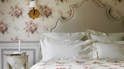 A neutral floral bedroom with statement headboard, white pillowcases, floral bedding, grey wall panelling, white and pink floral wallpaper, white nightstand and wooden sconce lamp with white lampshade