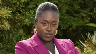 A posed character shot of Michelle Asante as Janelle Delice in Death in Paradise, wearing a magenta power suit and a white blouse underneath. She looks serious and unsmiling, and there is a lot of green vegetation in the background behind her.