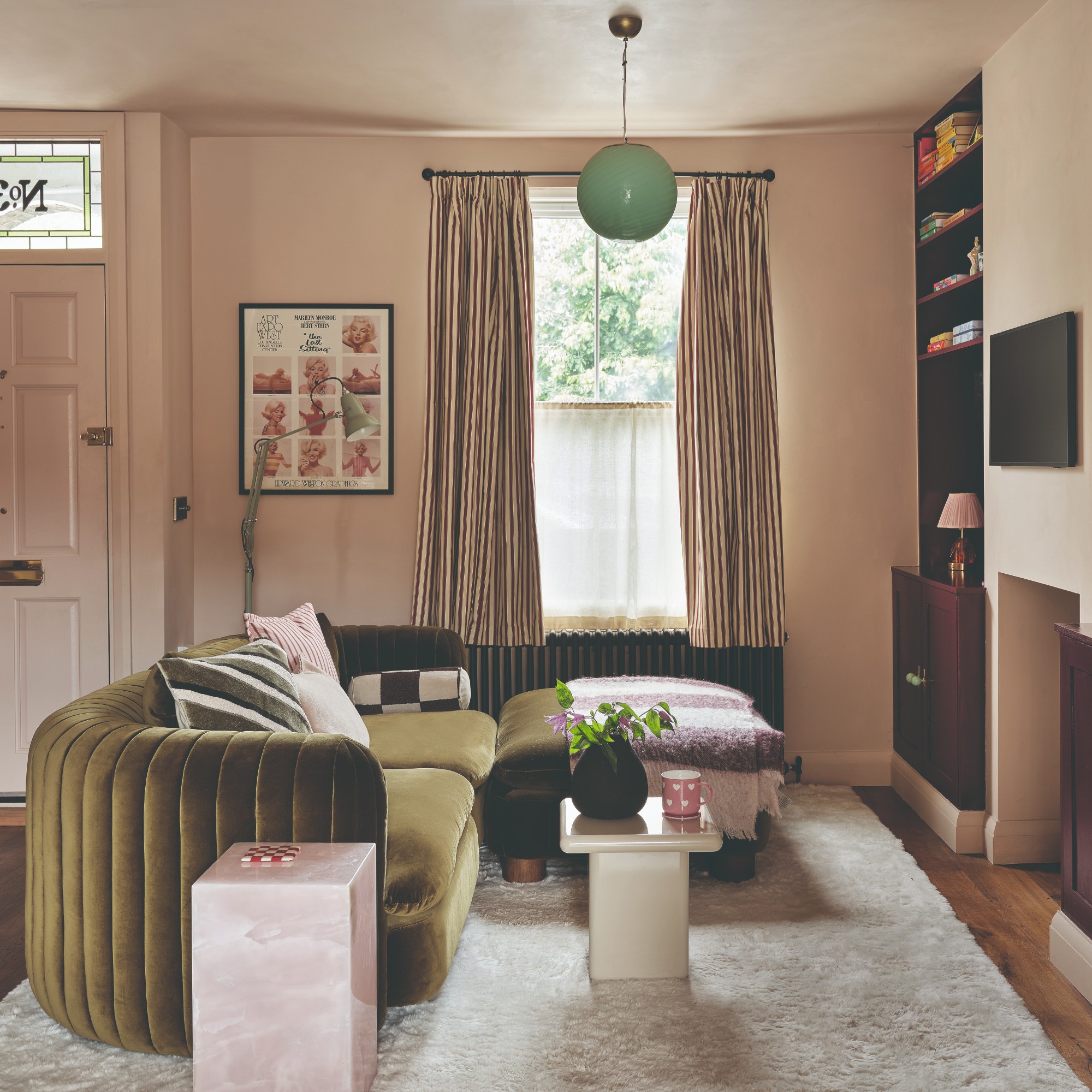 A small pink-painted living room with an olive green velvet curved sofa and burgundy bookcases built into alcoves