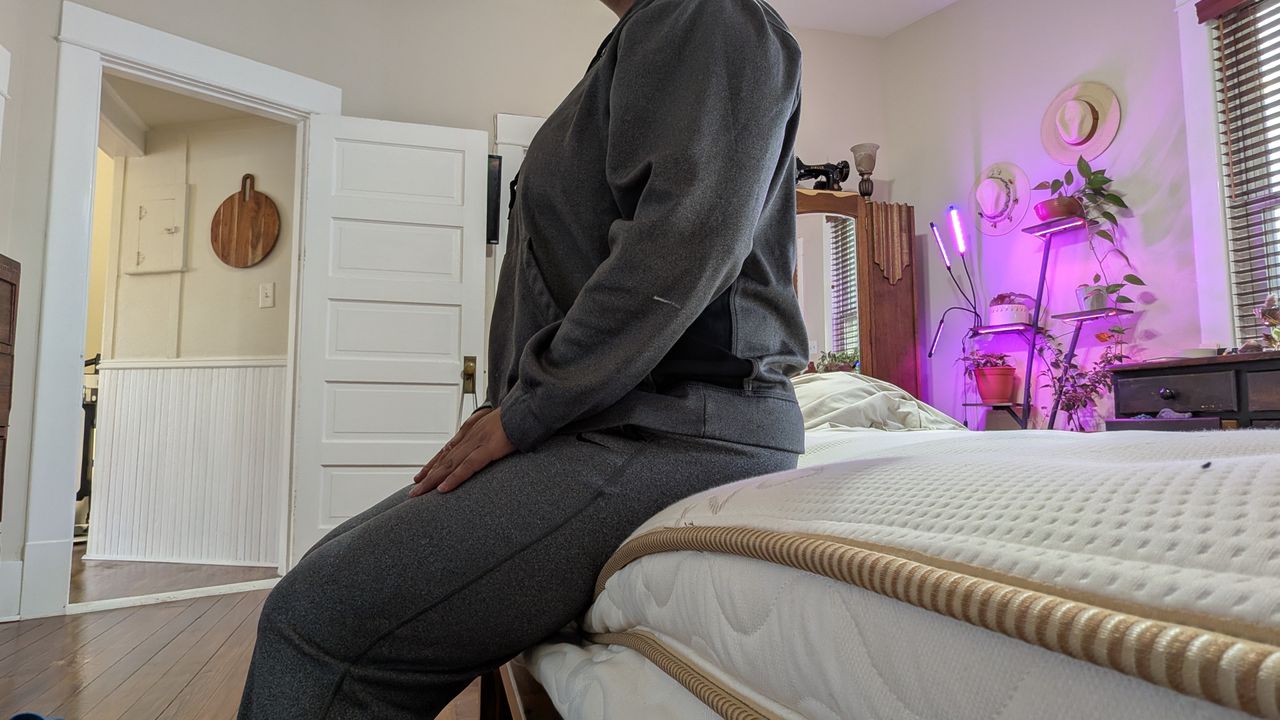 Tester Alex sitting on the edge of the Saatva Latex Hybrid Mattress in her bedroom.