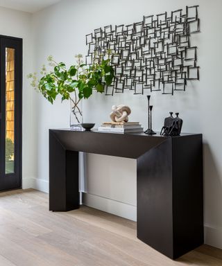 Modern entryway with large black metal console table, decorated with ornaments and vase, metallic geometric artwork on wall above, white painted walls, front door in corner