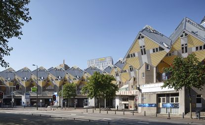 Exterior view of Rotterdam's Cube Houses during the day. The houses are yellow and grey cubes that are tilted 45 degrees. There are trees and a road outside