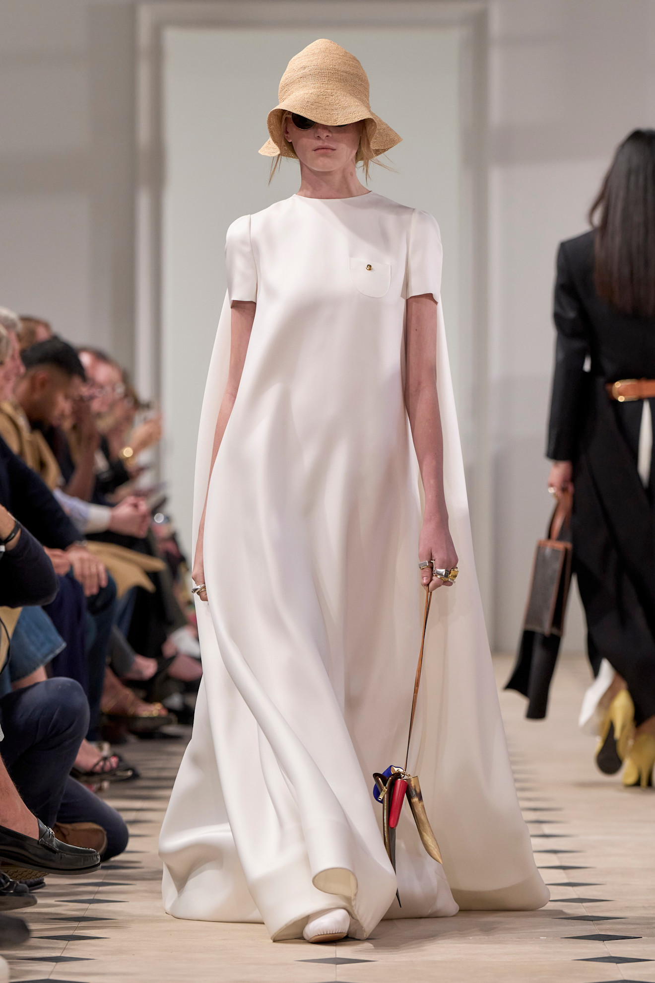 A model wears a white flowing maxi dress, a natural-toned woven straw hat, round sunglasses and white leather shoes on the Celine Ready to Wear Spring Summer 2026 runway.