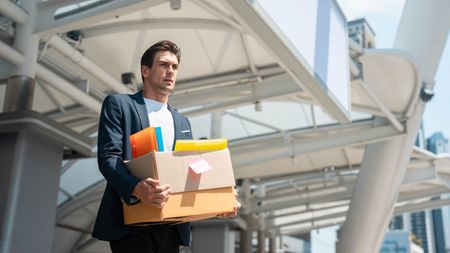 A man carries a box with belongings from his desk as he walks out of an office building.