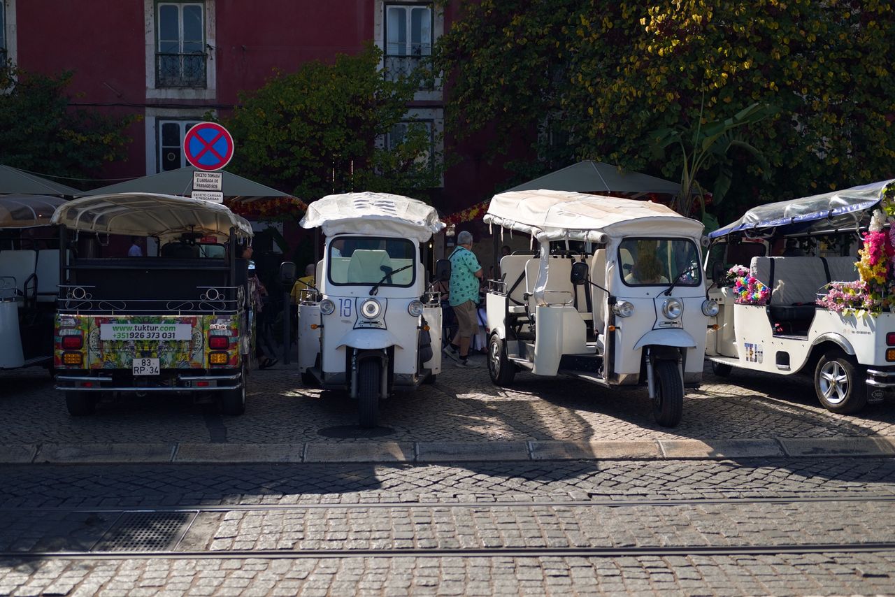 Row of parked tuk-tuks beside tram tracks on a cobbled Lisbon street, with leafy trees and a red building behind.