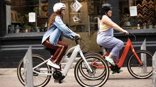 Two women - one white and one east Asian, cycling electric hybrid bikes together in an urban area, wearing casual clothing and helmets