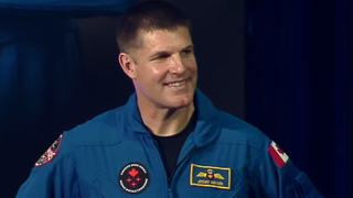 jeremy hansen smiling in his flight suit. on the front is a canadian space agency logo and his name tag. a canadian flag is on his left shoulder