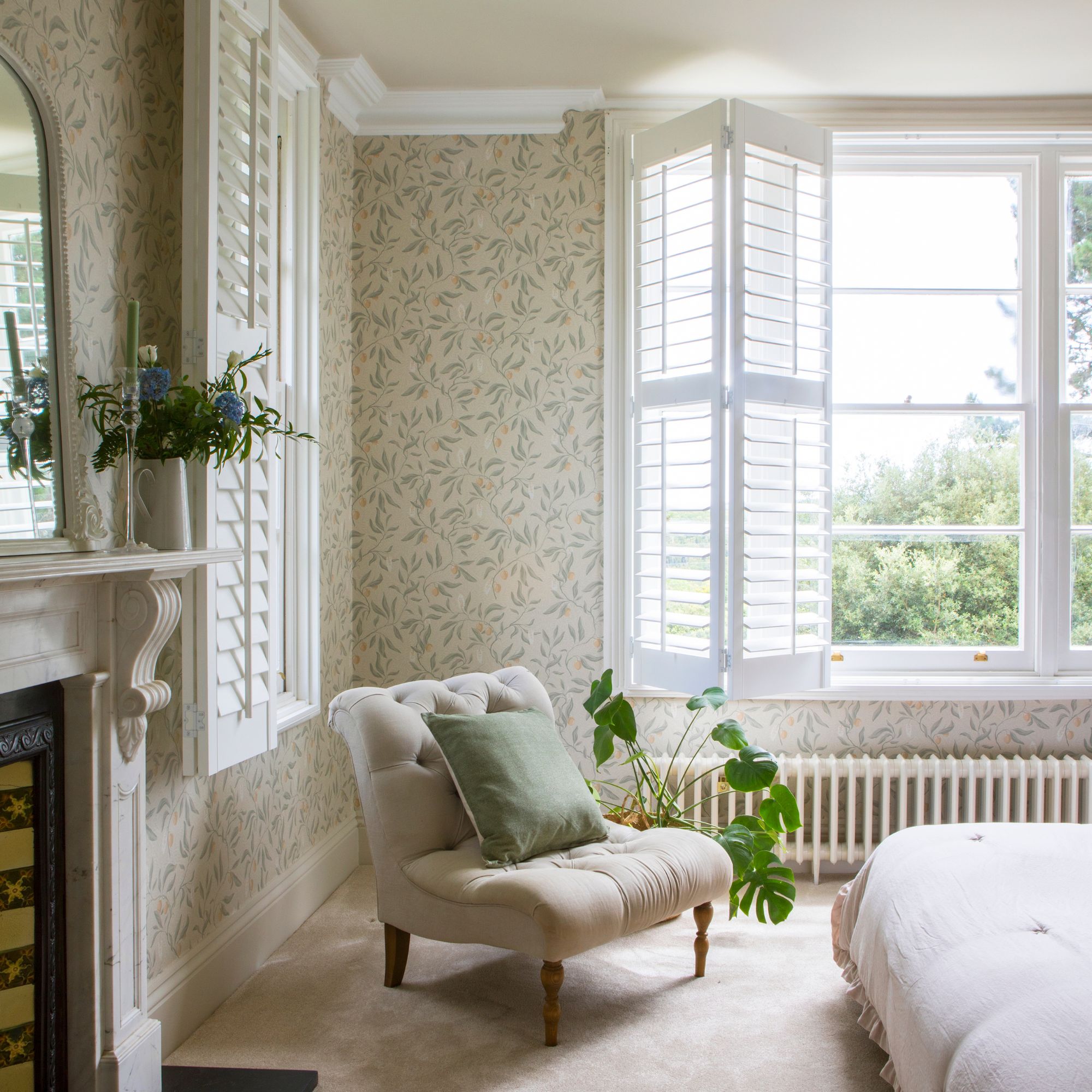A neutral, William & Morris wallpapered bedroom with large white shutters, cream carpet and a cream chair with a green cushion on.