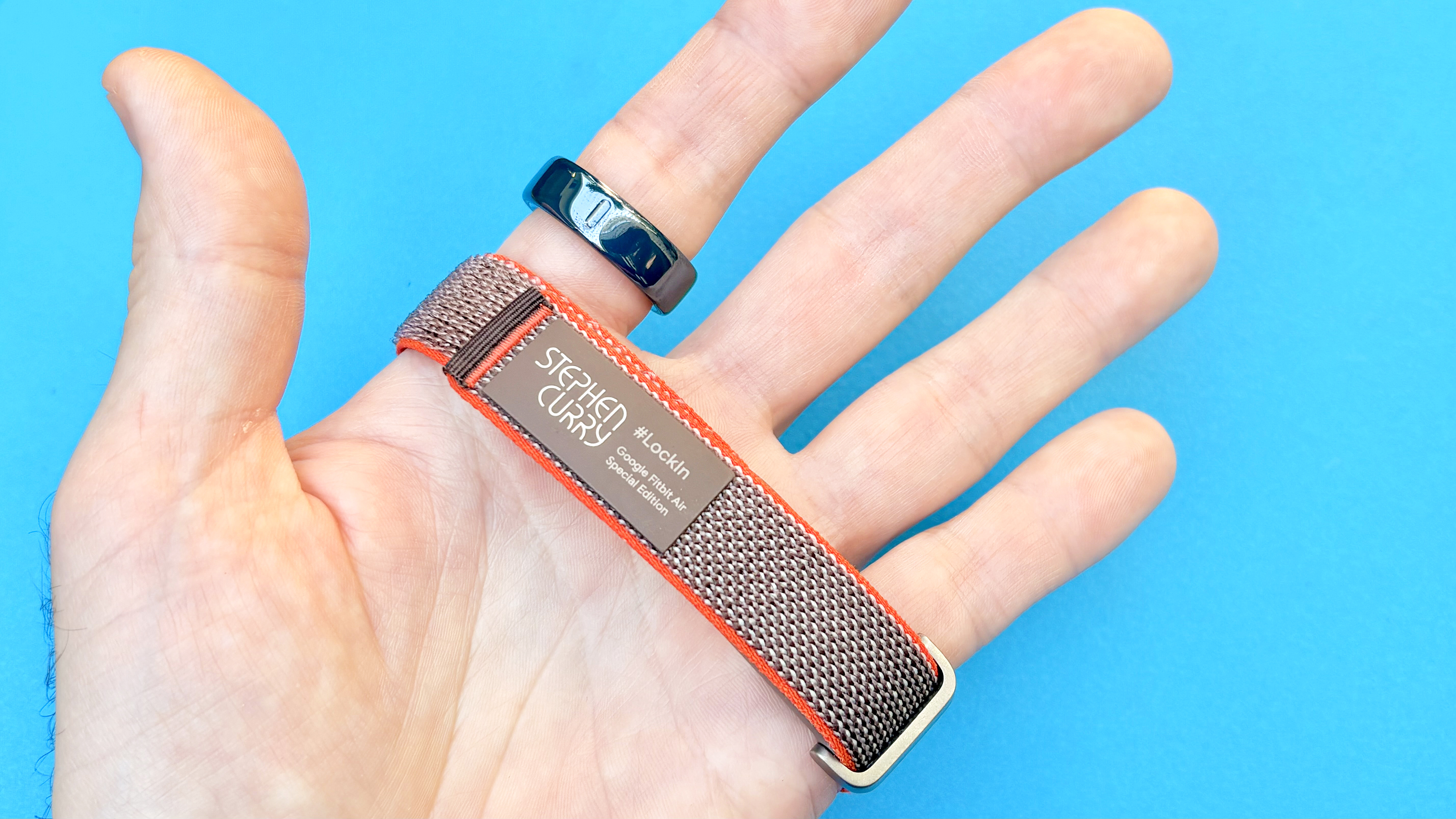 Close-up of the Fitbit Air and Oura Ring 4 Ceramic in the writer's hand against a blue background