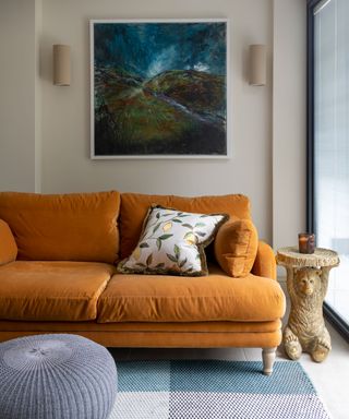 Living room with cozy orange sofa, artwork on wall behind, side table, round ottoman, blue square patterned rug