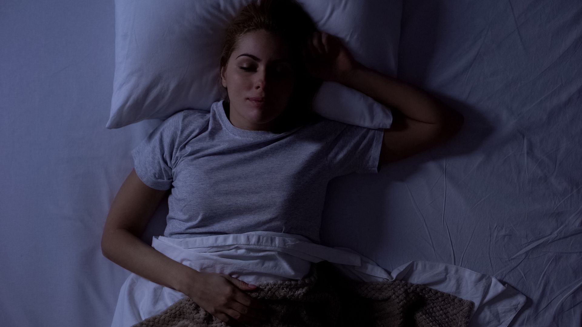 A woman lies in bed on her back in the dark with her eyes closed and one hand behind her head.